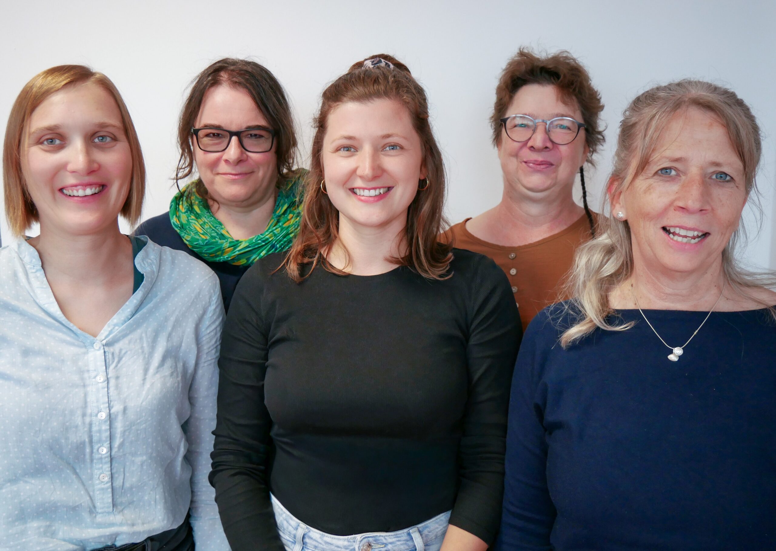 Fünf Frauen stehen dicht beieinander in einem Innenraum und lächeln in die Kamera vor einer schlichten weißen Wand. Sie tragen legere Oberteile und haben unterschiedliche Haarfarben und Frisuren.